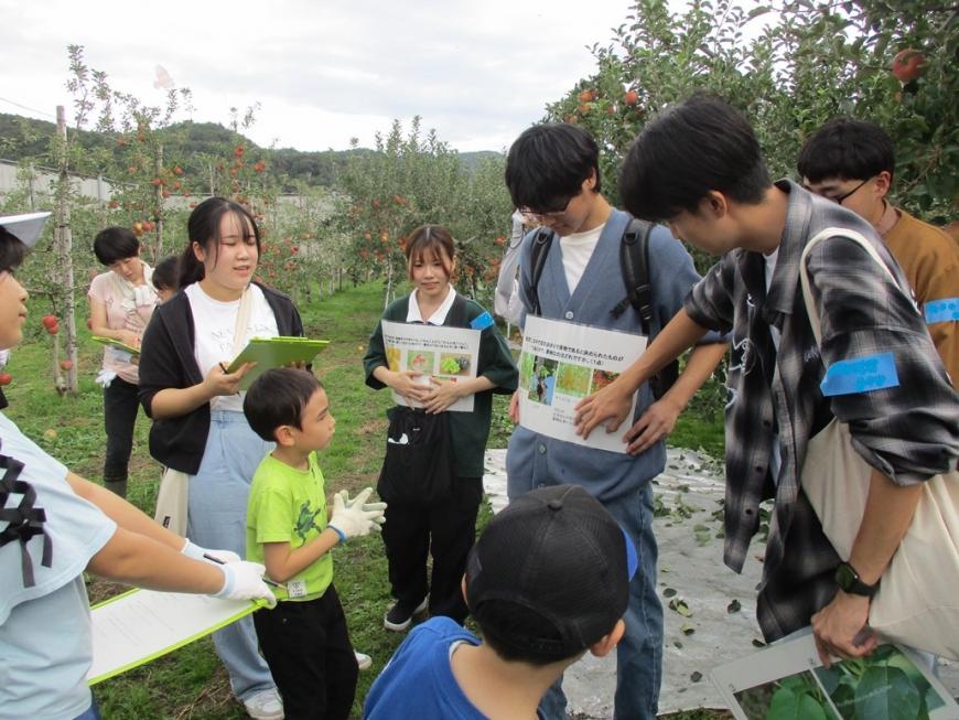 クイズが記載された紙を胸の前で持っている大学生の前に子どもたちが集まっている写真