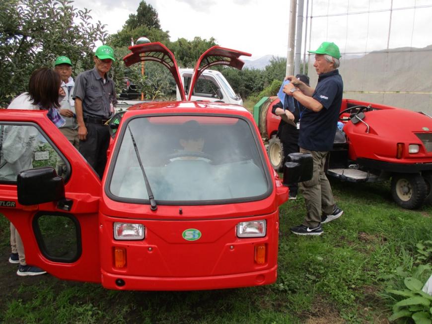 運転席がキャビンになっているタイプの赤いスピードスプレーヤーの周りに緑の帽子をかぶった男性たちと参加者がいる写真