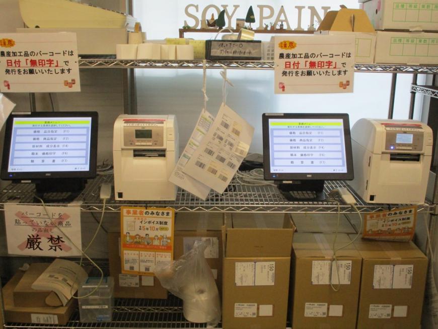 ラックにラベルを作成する機械が2台並べて置かれ、下の段に段ボール箱などが置かれている写真