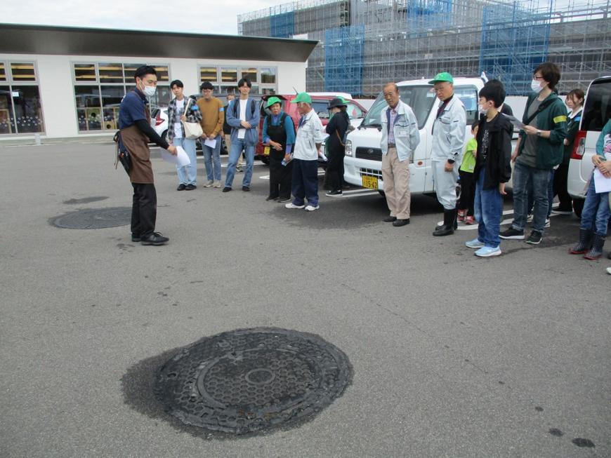 マンホールの手前で一列になり茶色いエプロンをつけた男性の説明を聞いている参加者たちの写真