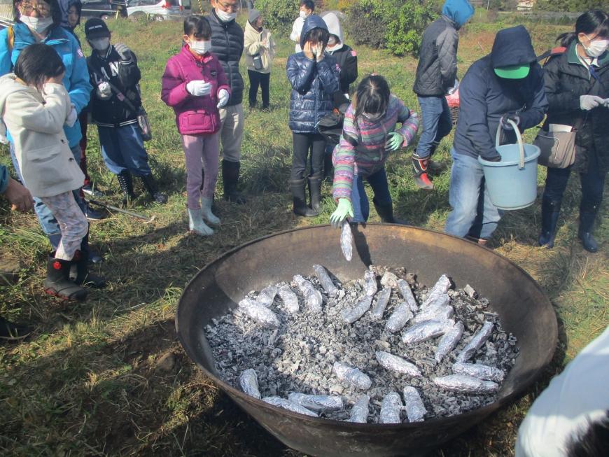 アルミホイルで巻かれたサツマイモが大きな炭化器にたくさん投入されており、周りで参加者たちが様子を見ている写真