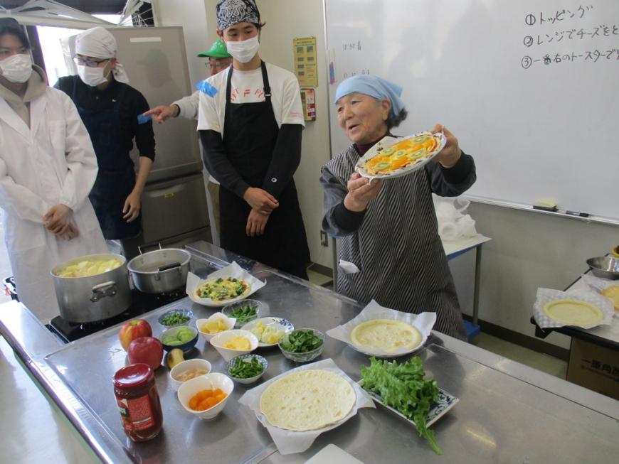 たくさんの材料が乗った調理台の前で、三角巾をつけた女性が見本のピザを参加者に見せている写真