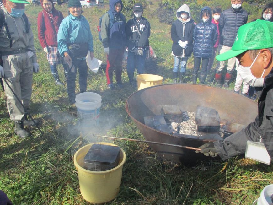 男性が長い棒に引っ掛けた缶を水を入れた黄色いバケツに着けている様子を炭化器の周りに集まった参加者たちが見守っている写真