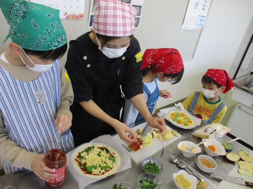 調理台で、参加者の女性や子供たちがフルーツピザと野菜ピザを作っている様子の写真