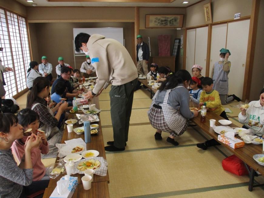 和室に並んだ2列の長机の席に座った参加者たちが、出来上がった料理などを食べている様子の写真