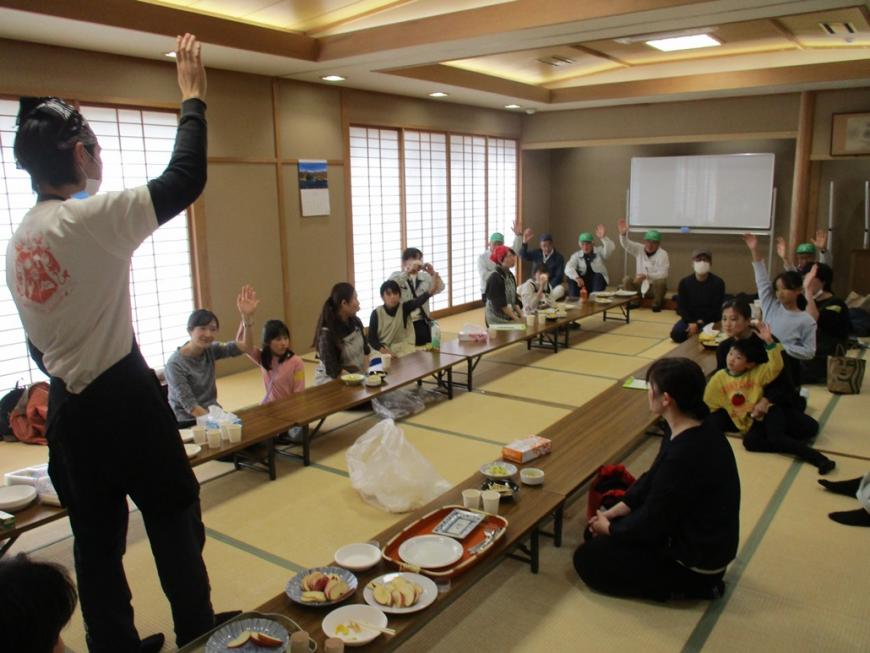 長机の間に立った男性が右手を挙げ、席に座った参加者たちの多数が手を挙げている様子の写真