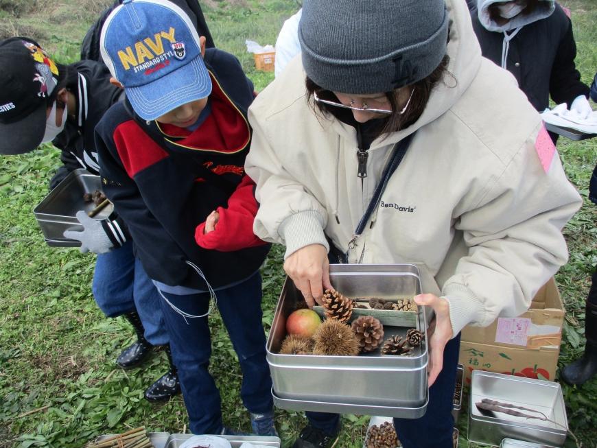 女性が持った缶に、松ぼっくり、イガグリ、竹、クルミ、りんごなどの材料が入っており、後ろから子どもがのぞき込んでいる様子の写真