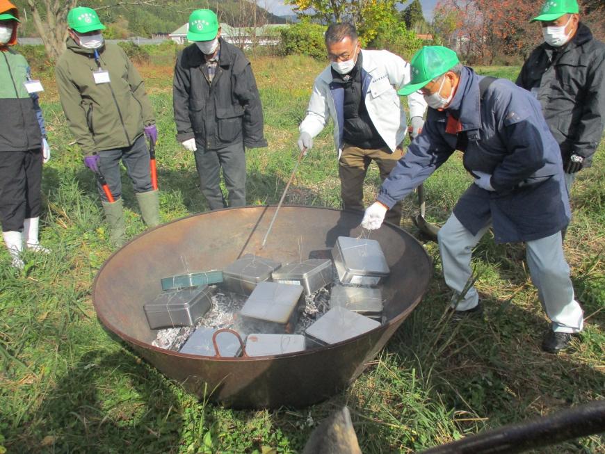 大きな炭化器にたくさんの四角い缶が投入され、棒を持った男性二人が位置を調節し、緑色の帽子をかぶった男性たちがその周りで様子を見ている写真