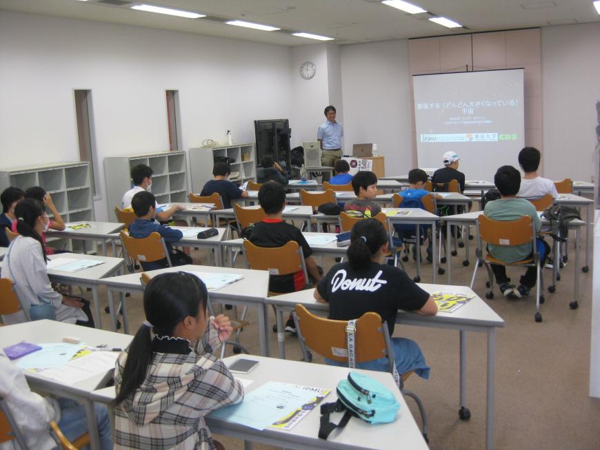 前方のスクリーンに資料が映され、その横に高田昌広教授が立っており、参加者の子ども達が長机の席について話を聞いている講座の様子の写真