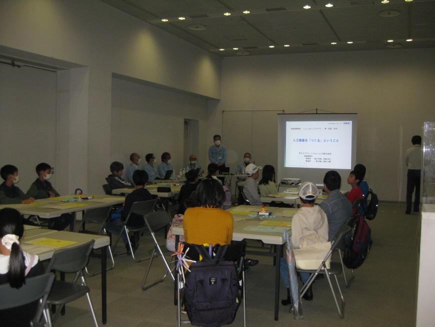 室内の前方に資料が映されたスクリーンが設置され、スクリーンの左側に関係者の男子が立っており、参加者の方々がテーブルに座って話を聞いている様子の写真