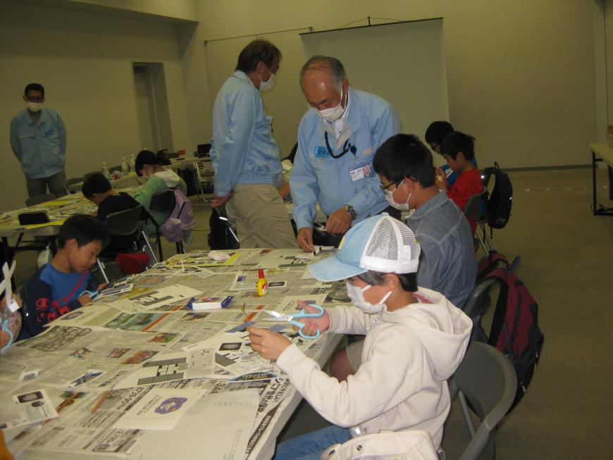 参加者の子ども達ががハサミで紙を切り抜く作業をおこなっており、関係者の男性が側で作業を手伝っている様子の写真