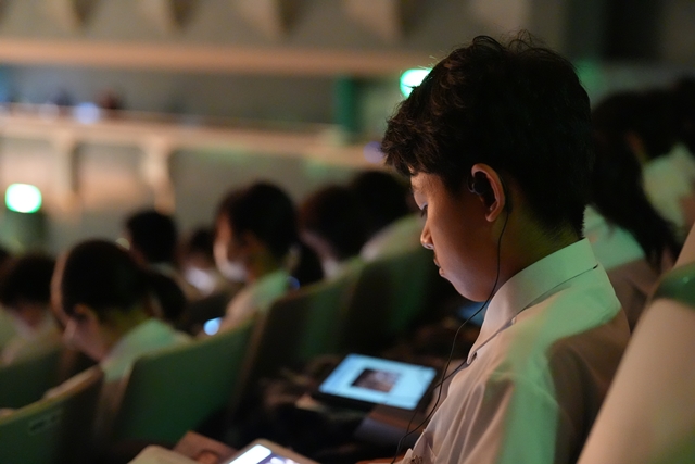 客席で、イヤホンを耳につけ、タブレット端末の映像を見ている男子生徒を写した写真