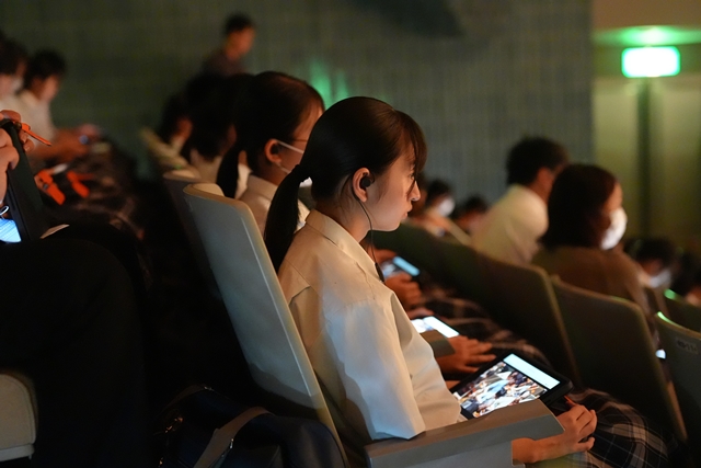 客席でイヤホンを耳につけ、膝の上には映像が映されたタブレット端末が置かれ、舞台に注目している女子生徒を写した写真