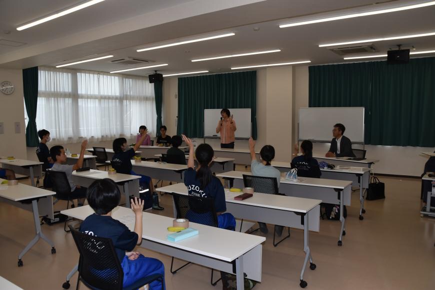 美術部員の皆さんが席について手をあげており、齋氏が前に立ちマイクを持って話をしている様子の写真