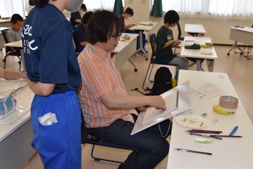齋氏が生徒の席に座り、画用紙にデッサンしており、すぐ横に生徒が立って、デッサンしている様子を見ている写真