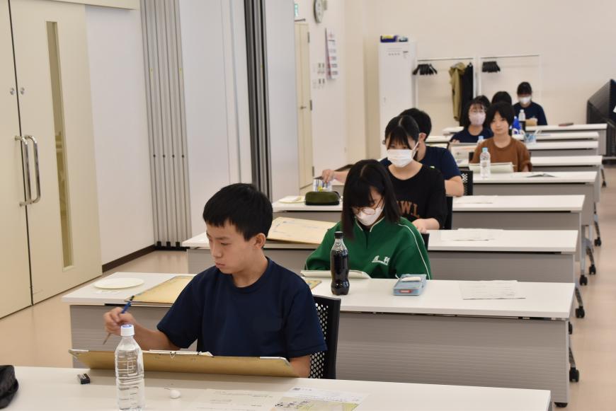 長机が教室の後ろまで並んでおり、生徒がそれぞれ座って、机の上に置かれた飲み物が入ったペットボトルをデッサンしている様子の写真