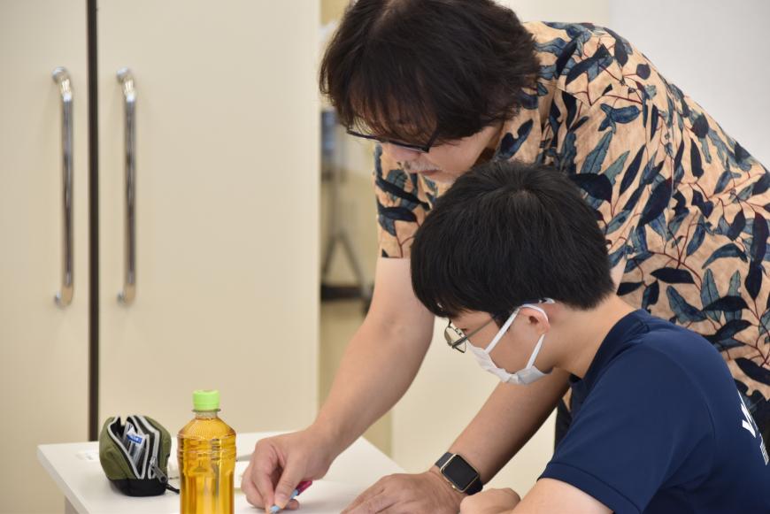 齋氏が男子生徒の机で実際に鉛筆を握り画用紙に直接書き込み、男子生徒が真剣な表情で指導を受けている様子の写真