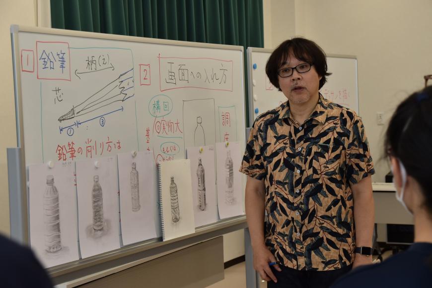生徒の作品がホワイトボートに並んでおり、齋氏が生徒の方を見てコメントをしている様子の写真
