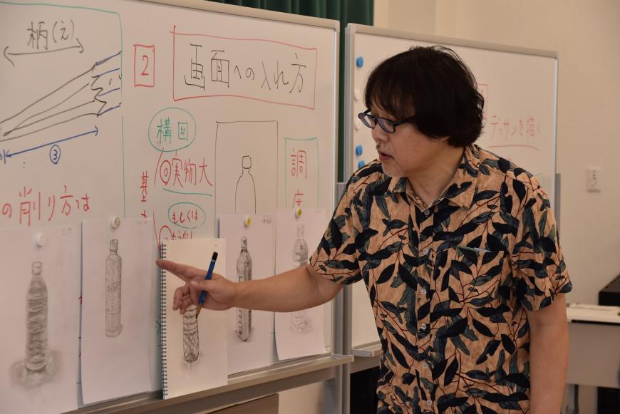 ホワイトボートに生徒の作品が並べて掲示されており、齋氏が作品の一つを指さししながらアドバイスをしている様子の写真
