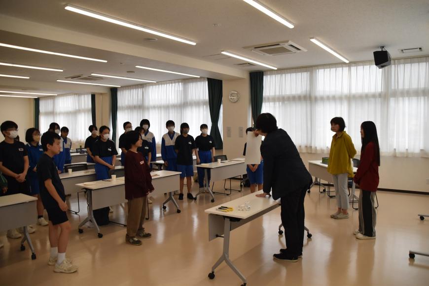 齋氏が画用紙を持って生徒に説明をしており、多くの参加者の生徒が立って齋氏の話を注目して聞いている様子を写した写真