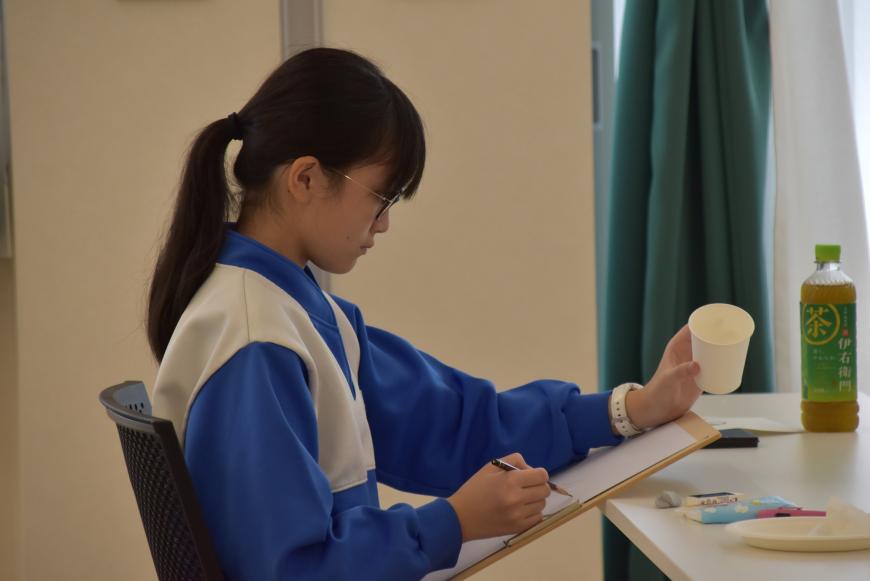 左手で紙コップを握り、右手で画用紙にデッサンしている女子生徒を写した写真