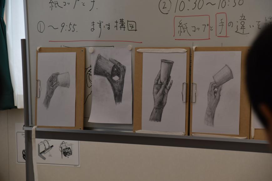 色々な方向から描かれた紙コップを持った手のデッサン4枚の生徒の作品が並んでいる様子の写真