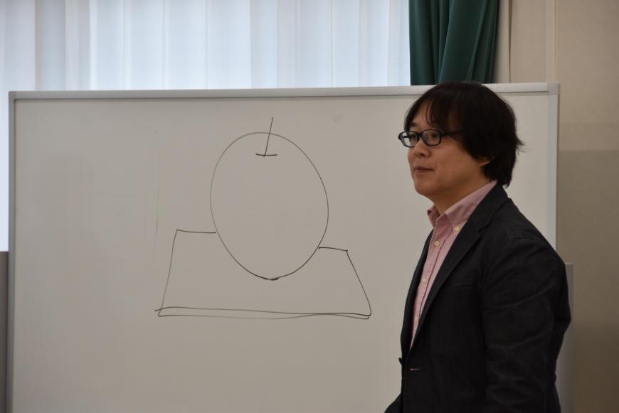 白版にリンゴの絵が描かれ、齋氏が教壇に立っている写真