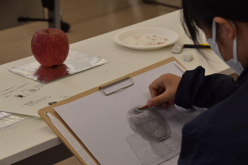 机の上のアルミホイルの上に置かれたリンゴを鉛筆で画用紙にリンゴの絵をデッサンしている生徒を左斜め後ろから写した写真