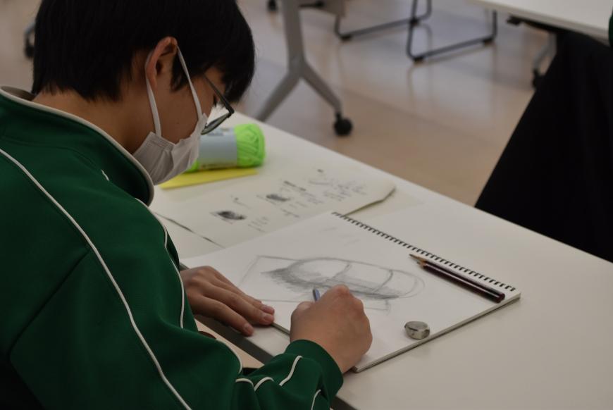 画用紙に折り紙の上に置かれた毛糸玉のデッサンが描かれ、鉛筆で影の部分を作りだしている生徒の様子を写した写真