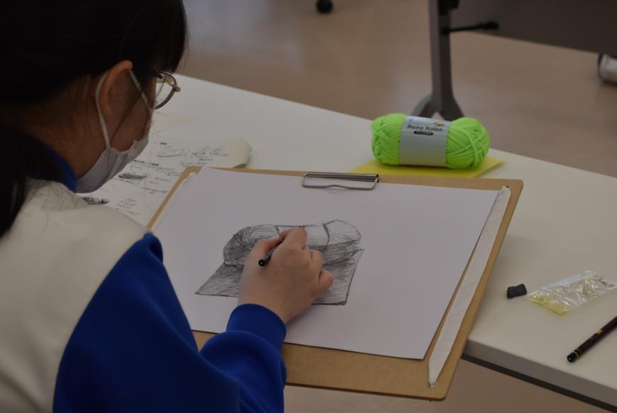 画用紙に折り紙の上の毛糸玉が描かれ、毛糸の質感を描き加えている生徒の様子を写した写真