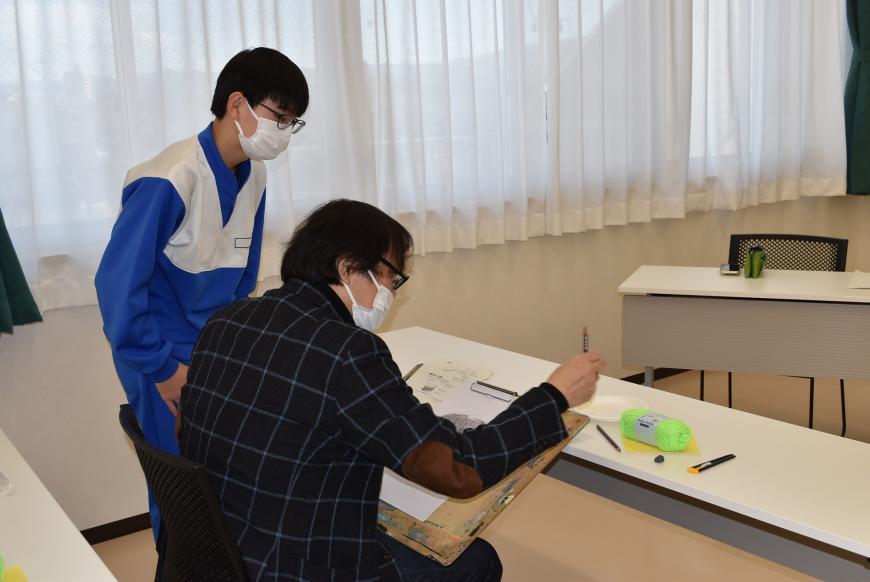 生徒の席に齋氏が座り、画用紙に描きながら直接指導をしており、側で男子生徒が真剣に見ている様子の写真