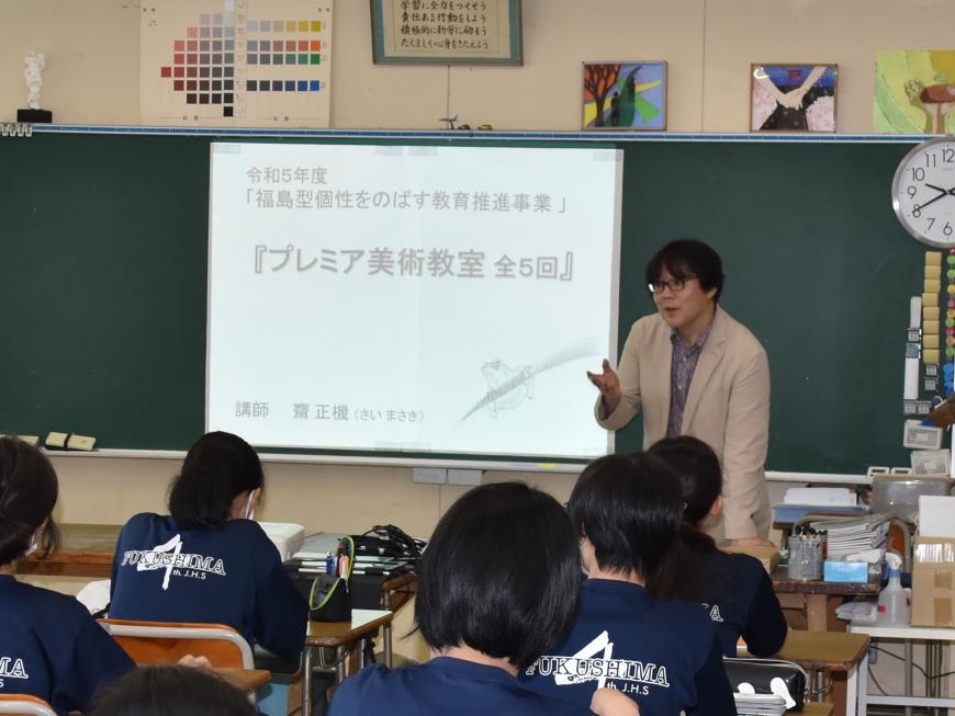 プレミア美術教室 全5回と教室の黒板のスクリーンに映像が映され、齋氏が教壇で生徒の皆さんに話をしている様子の写真