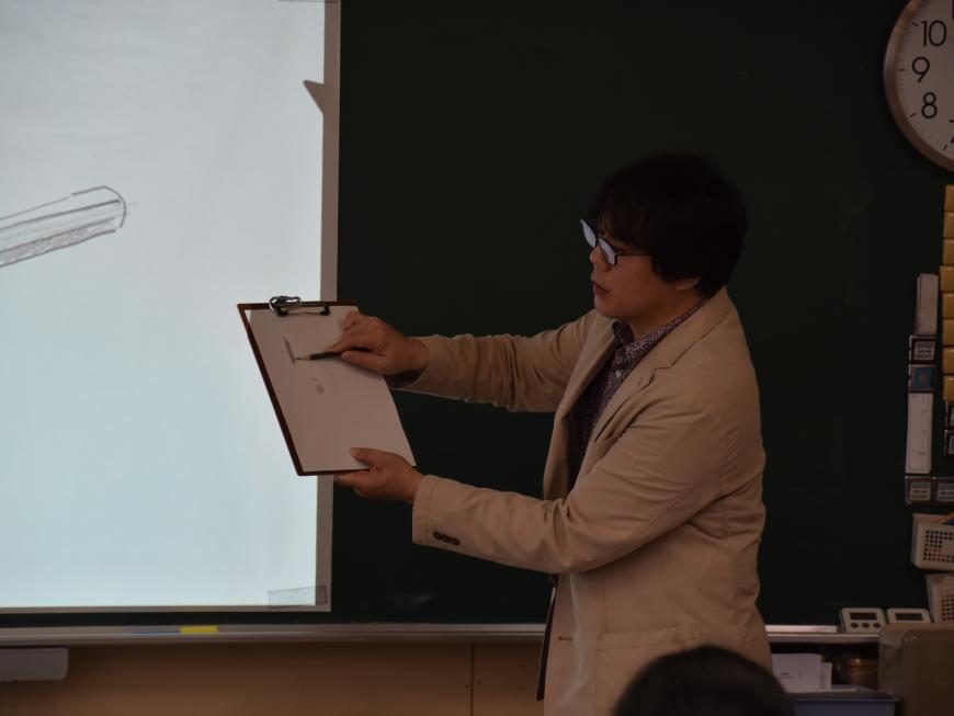 齋氏が鉛筆で画用紙にデッサンをしながら描き方の説明をしている様子の写真