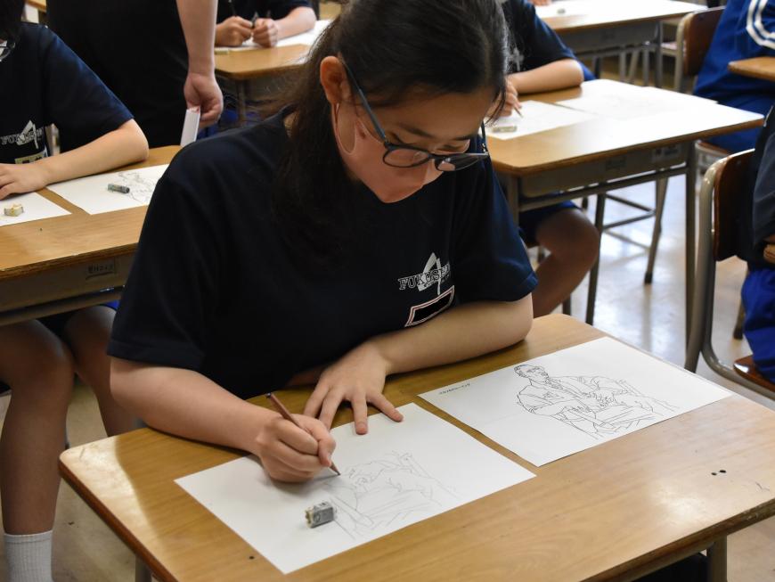 机の上に置かれた見本の絵を見ながら、女子生徒が画用紙にデッサンしている様子を写した写真