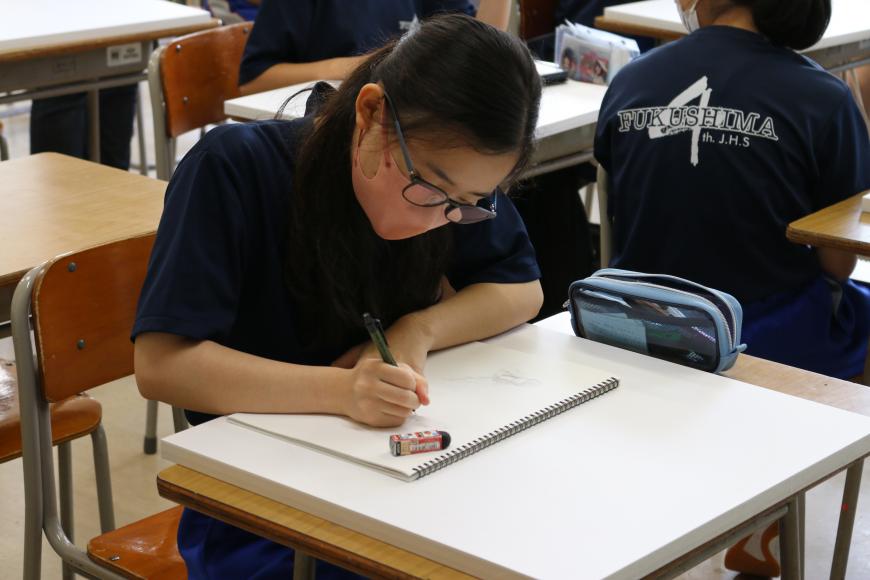 自席に座っている女子生徒が机の上に広げたスケッチブックに集中してデッサンをしている様子の写真