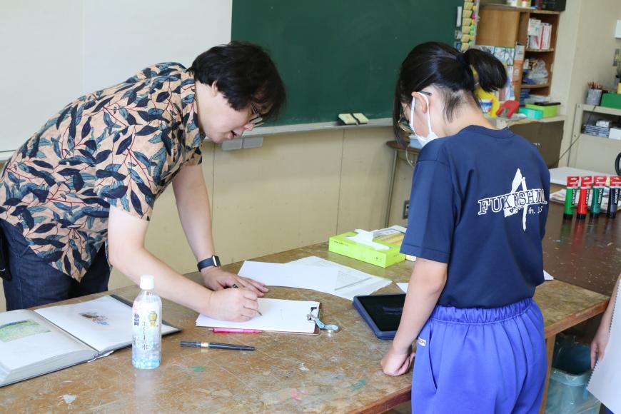 齋氏が紙に書き込みながら一人の女子生徒に説明をしている様子を写した写真