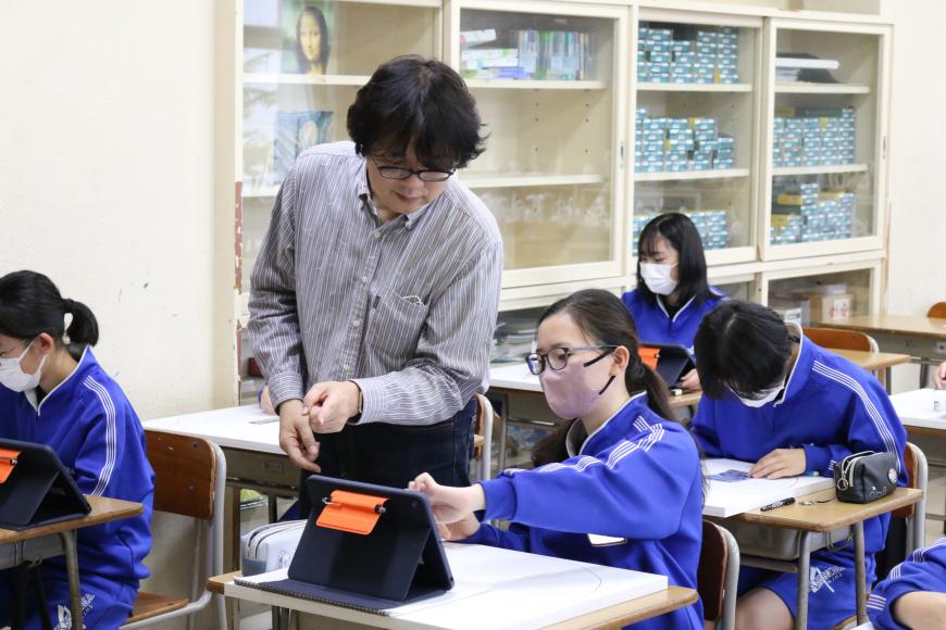 女子生徒の机の上に置かれたタブレットを女子生徒と傍に立つ齋氏が指さし覗き込んでいる授業の様子の写真