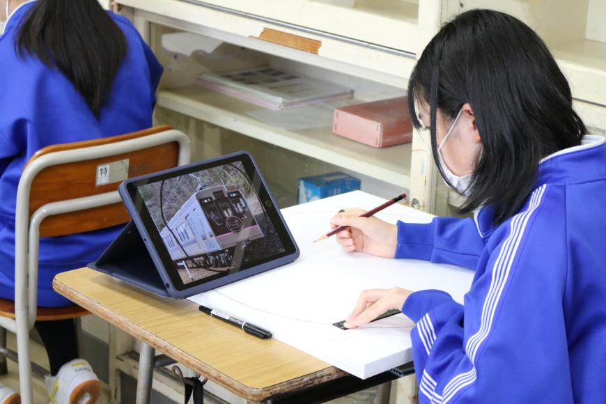タブレットに電車の映像が映され、女子生徒がタブレットを見ながら鉛筆で描いている様子の写真