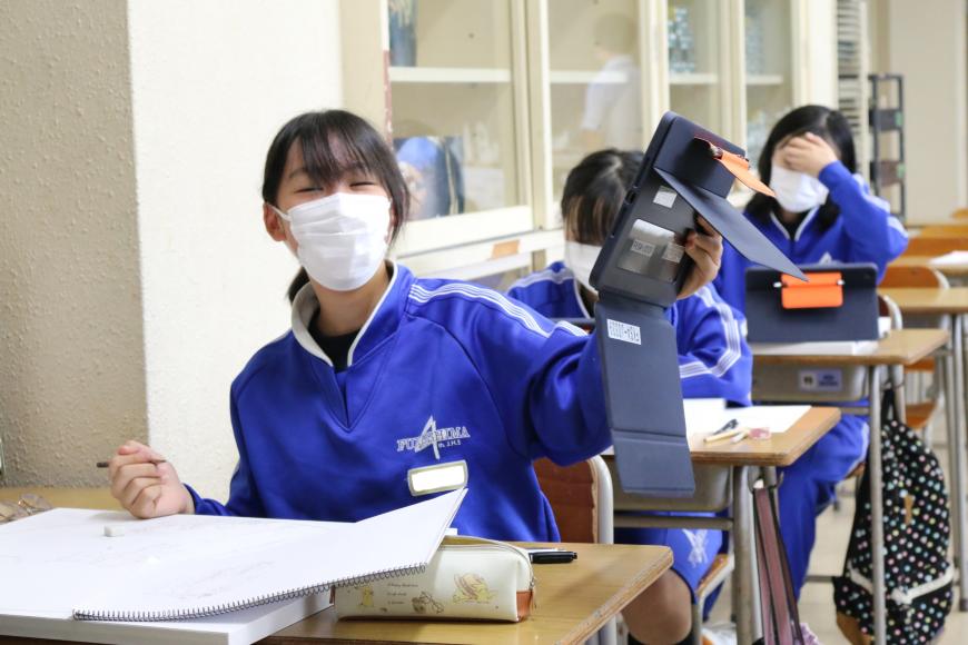 自席に座っている女子生徒がタブレット端末を左手に持ち、笑顔でカメラ目線で写っている写真