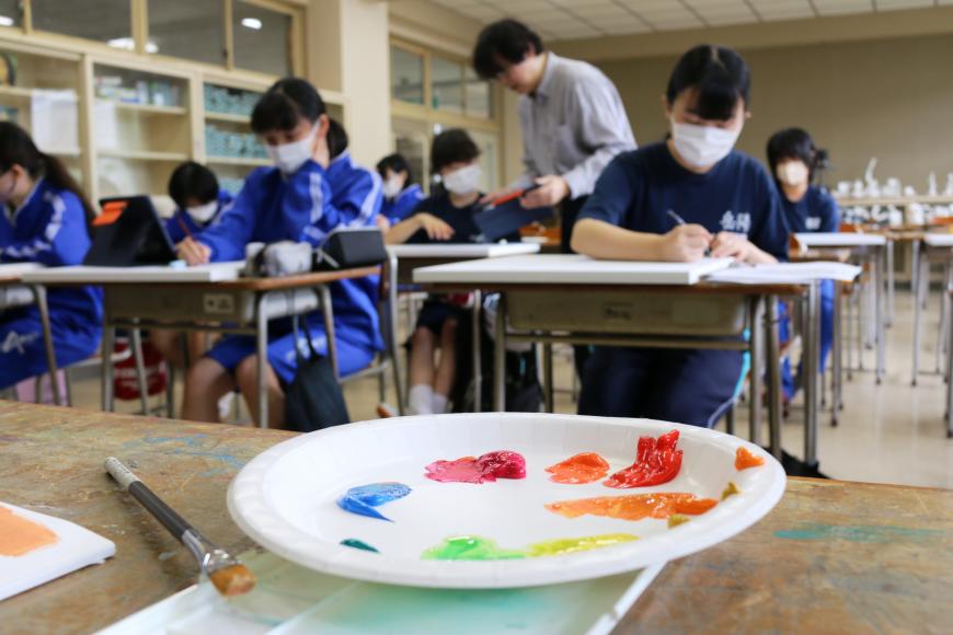 赤や青、オレンジ、黄色、緑などの絵具が出された紙皿をアップに、奥には生徒の皆さんが自席に座り鉛筆で集中して絵を描いている様子の写真
