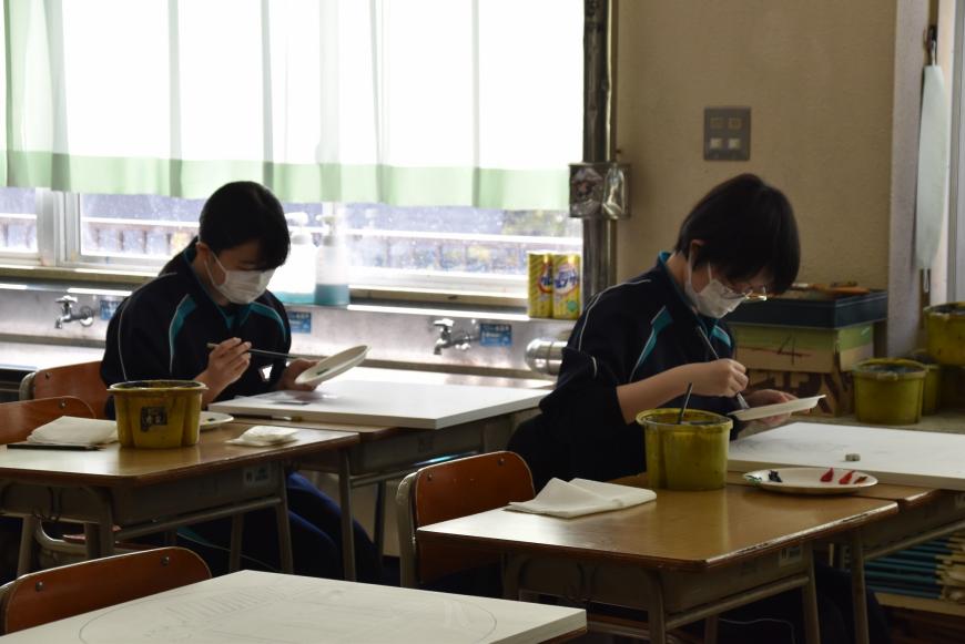 自席に座っている生徒が紙皿の絵具を筆に付けようとしている様子の写真