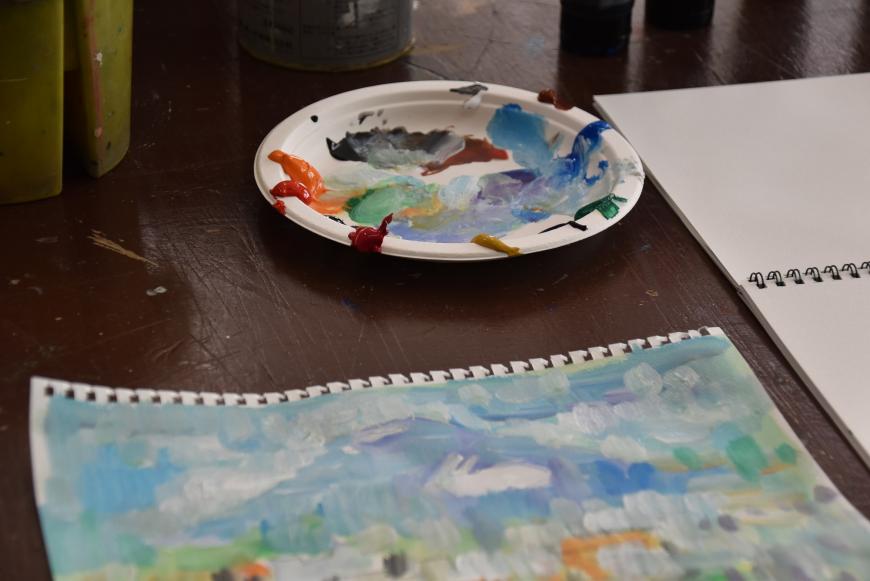 色々な色の絵具が出され使われて色が混ざっている紙皿と色々な色が塗られて山の絵が描かれた作品が置かれている写真
