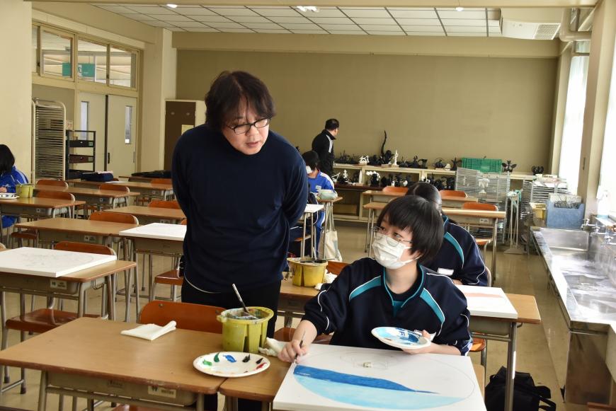 女子生徒が作品に青系の色を塗っており、生徒の側に立つ齋氏が生徒の絵を見ながら話しかけている様子の写真