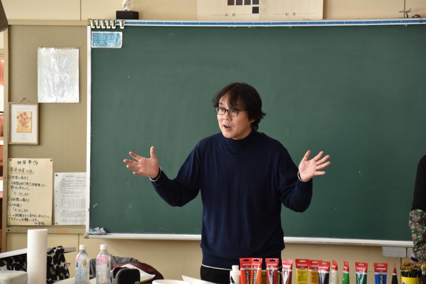 齋氏が黒板の前に立ち、両手を広げて話をしている様子の写真
