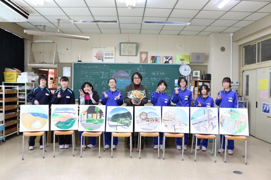 椅子の上に生徒の皆さんの作品が並べられ、その後ろに綺麗な花束を持った齋氏を中心に生徒の皆さんが並んで写っている記念写真