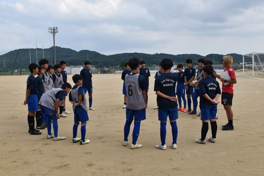 赤色のユニフォームを着た福島ユナイテッドFCの指導者の男性の周りに部員の皆さんが円になって集まり、話を聞いている様子の写真