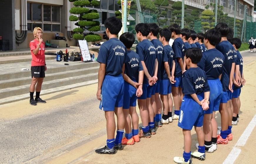 2列に整列した松陵中学校のサッカー部員の皆さんを前に、赤色のユニフォームを着た福島ユナイテッドFC指導者の男性が話をしている様子の写真