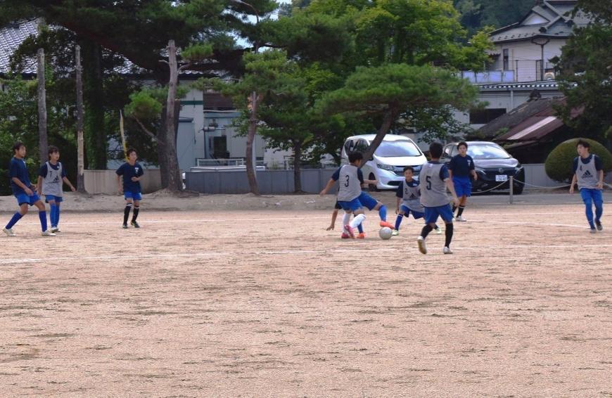 グラウンドで2チームに分かれた部員たちが、ボールを競り合いサッカーをしている様子の写真