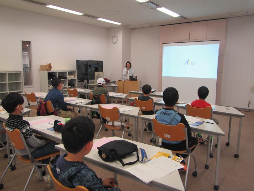 前方にスクリーンが設置され、その左側でマイクを持った講師の女性がマイクを持って話をし、参加者の子ども達が机の前の席に座って話を聞いている様子の写真