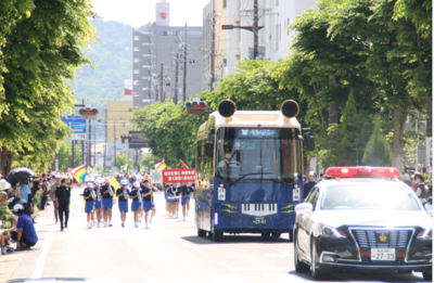 パトカー、青バスを先頭に生徒達が道路の中央を歩いて演奏している様子の写真